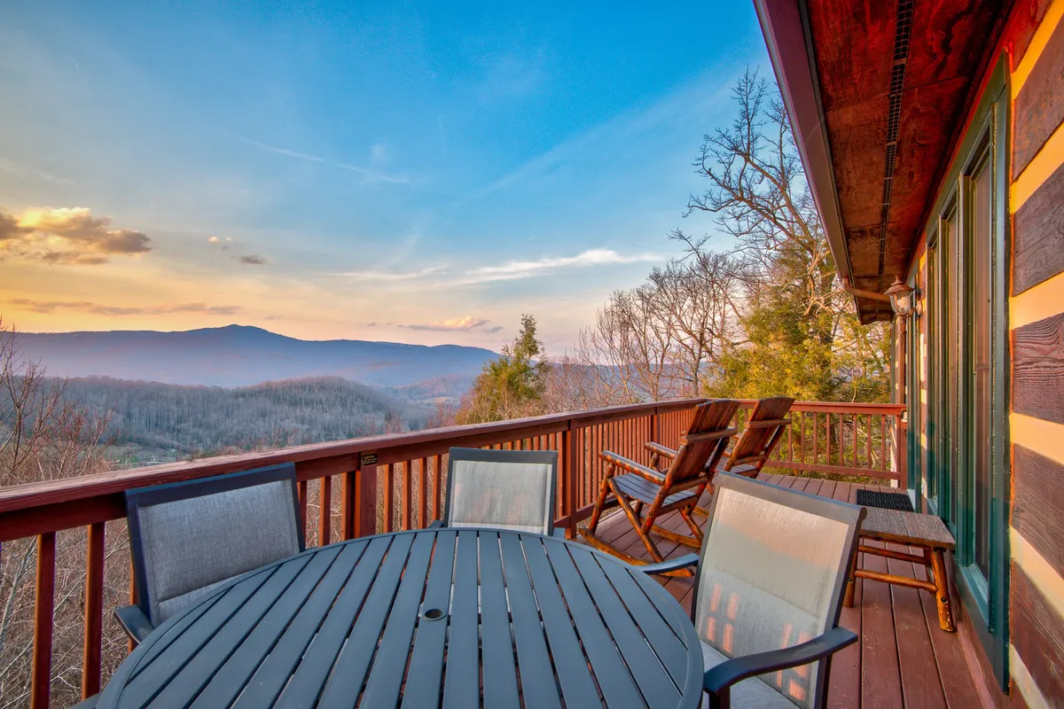 A spacious deck set against a backdrop of mountain views, featuring a dining table for al fresco meals and comfortable outdoor seating. The wooden deck is surrounded by a secure railing, and the tranquil ambiance is enhanced by the fading sunlight, perfect for enjoying sunsets or evening gatherings.
