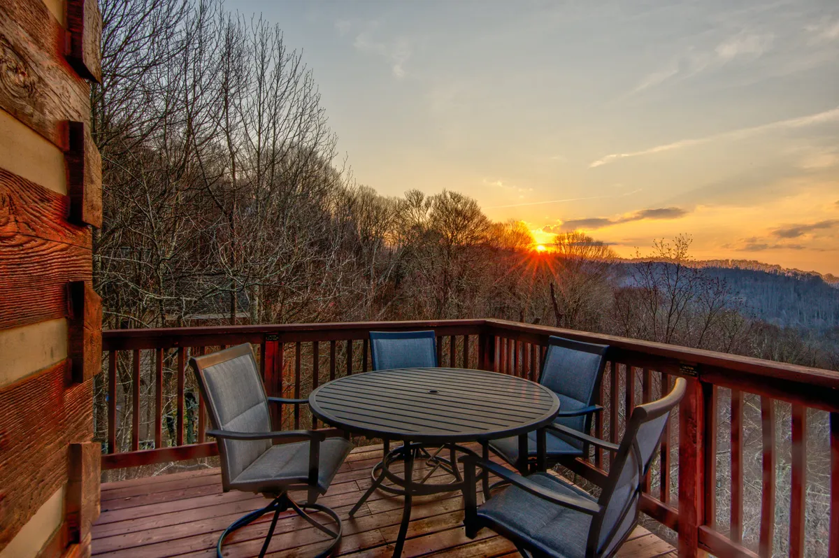 A wooden deck furnished with comfortable outdoor seating and a dining area, all facing a vivid sunset sky over the mountains. The space feels open, inviting, and secure, designed for both socializing and quiet reflection.