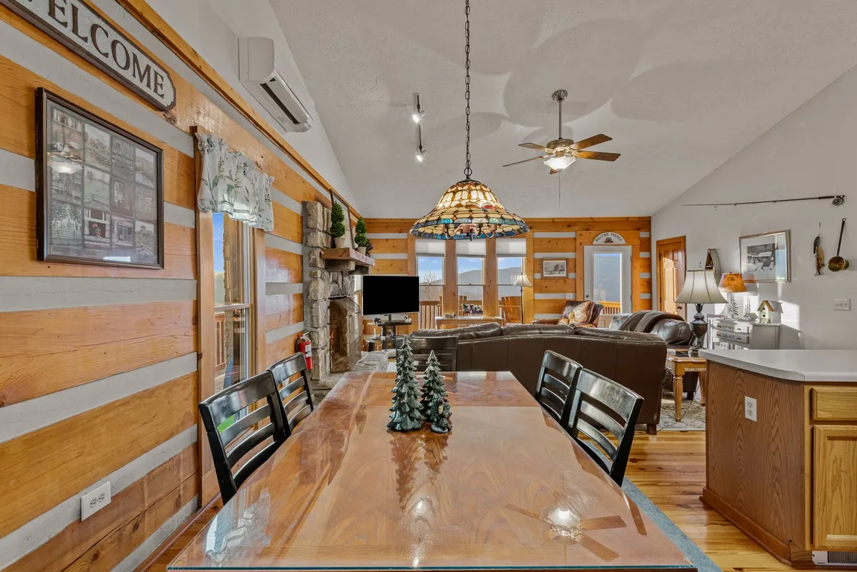 A spacious open-concept area that blends a wooden dining table, stained glass light fixture, stone fireplace, and comfortable seating. Large windows flood the room with natural light, revealing panoramic mountain scenery while setting the scene for shared meals and memorable gatherings.