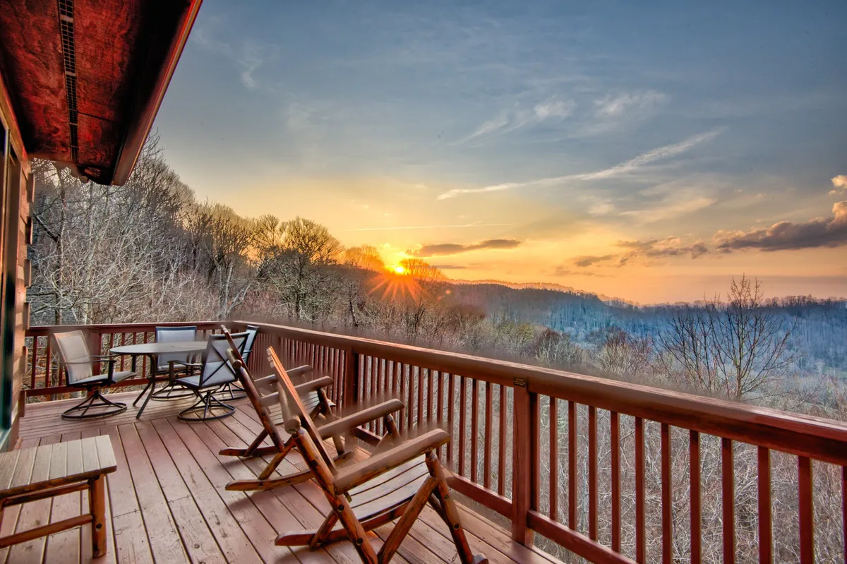 The image showcases the exterior of a cozy log cabin perched on Sugar Mountain at sunset. A spacious deck with comfortable outdoor furniture invites relaxation, while sweeping long-range mountain views unfold beyond. The sky is ablaze with vibrant sunset hues, promising tranquil evenings and the magic of stargazing after dark.
