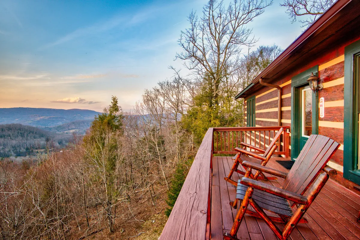 A cozy outdoor area with wooden rocking chairs arranged on a deck, overlooking breathtaking mountain vistas. The gentle glow of sunset bathes the scene, ideal for sipping coffee or simply relaxing while soaking in the serene mountain landscape.