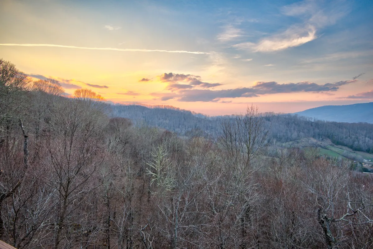 A sweeping view of the sunset sky over a mountain range, framed by bare trees, evoking the majesty of the natural landscape. The open horizon suggests possibilities for hiking, photography, and wildlife spotting.