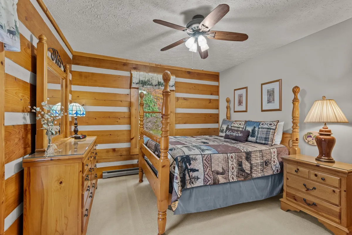 A cozy queen bedroom featuring a wooden bed frame set against a striped wooden wall, a soft bedside lamp, and a dresser with a mirror. Sunlight streams in from the window, creating a welcoming and tranquil environment for relaxation.