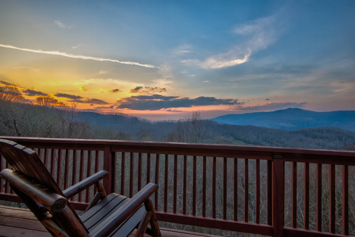 This image captures a classic mountain view at sunset, with a cozy rocking chair set on a deck overlooking a stunning mountain range. The soft evening light and tranquil atmosphere suggest an ideal spot for couples to relax, converse, and enjoy each other's company while taking in the dramatic natural scenery.