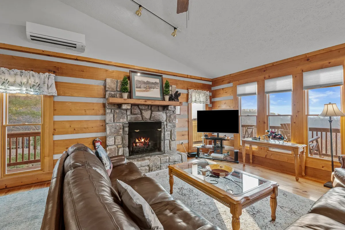 This image features a warm and welcoming great room showcasing a stone fireplace, comfortable leather sofa, a coffee table, and large windows that reveal stunning mountain views. The space is filled with natural light and is perfect for relaxing, socializing, or unwinding by the fire.