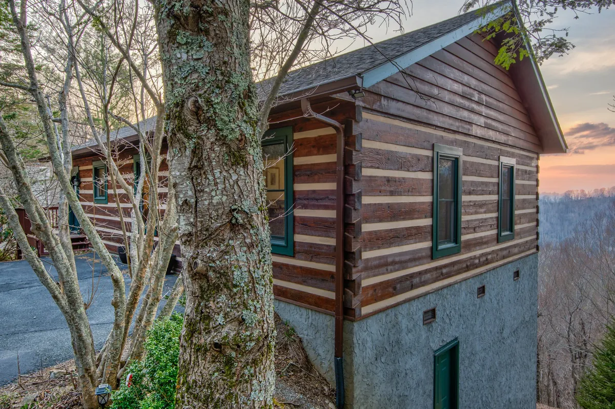 A log cabin nestled among mature trees, with the sun setting in the background. The spacious porch and surrounding natural landscape create a serene, inviting atmosphere that emphasizes outdoor relaxation and mountain living.