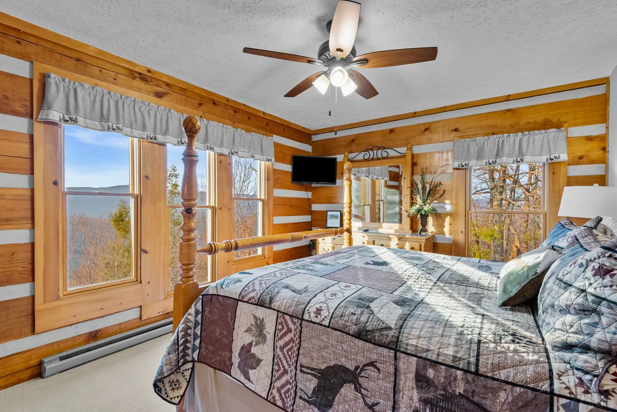 A serene primary bedroom featuring a wooden bed frame with a plush patchwork quilt, large windows framing scenic mountain views, a wooden dresser, and a ceiling fan overhead. The cozy atmosphere is enhanced by gentle natural light, making this a perfect retreat for restful nights.